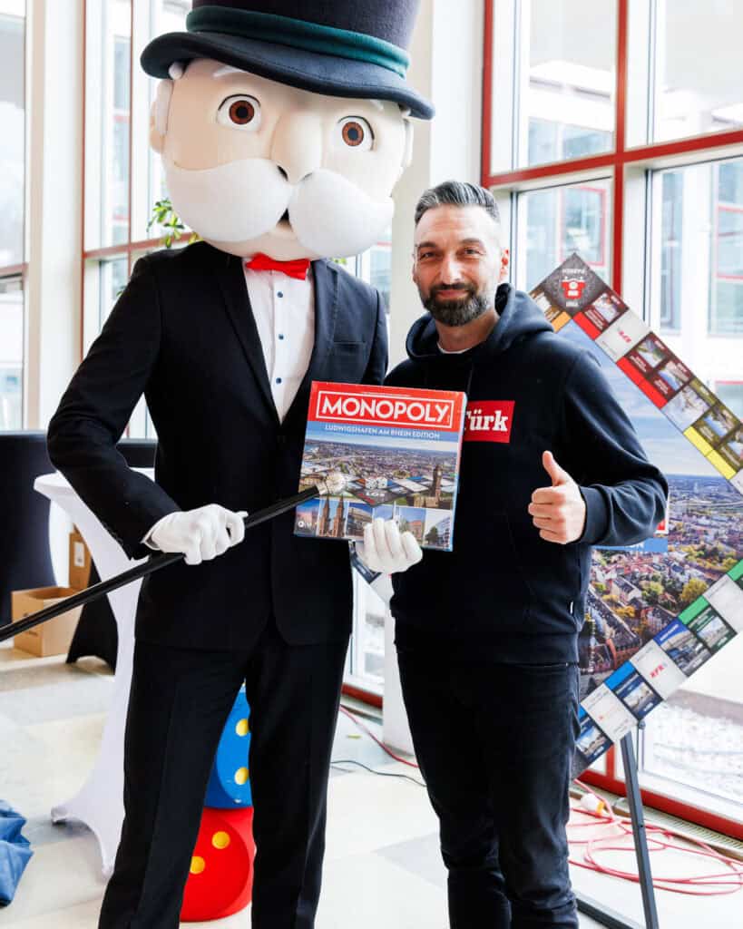 Pälzer Türk hält ein Monopoly Spiel in der Hand und steht neben der Monopoly Figur