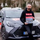 Pälzer Türk steht vor dem Auto mit dem Monopoly Spiel in der Hand und lacht