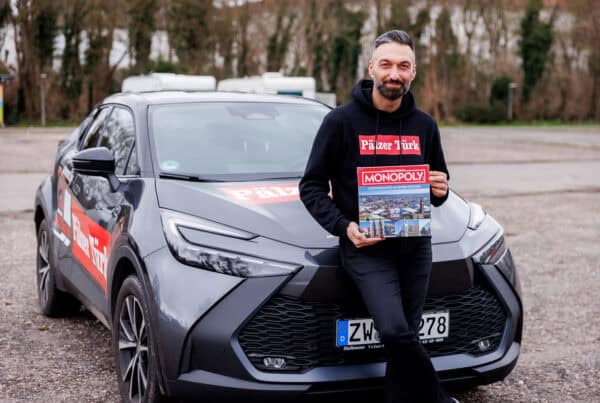 Pälzer Türk steht vor dem Auto mit dem Monopoly Spiel in der Hand und lacht